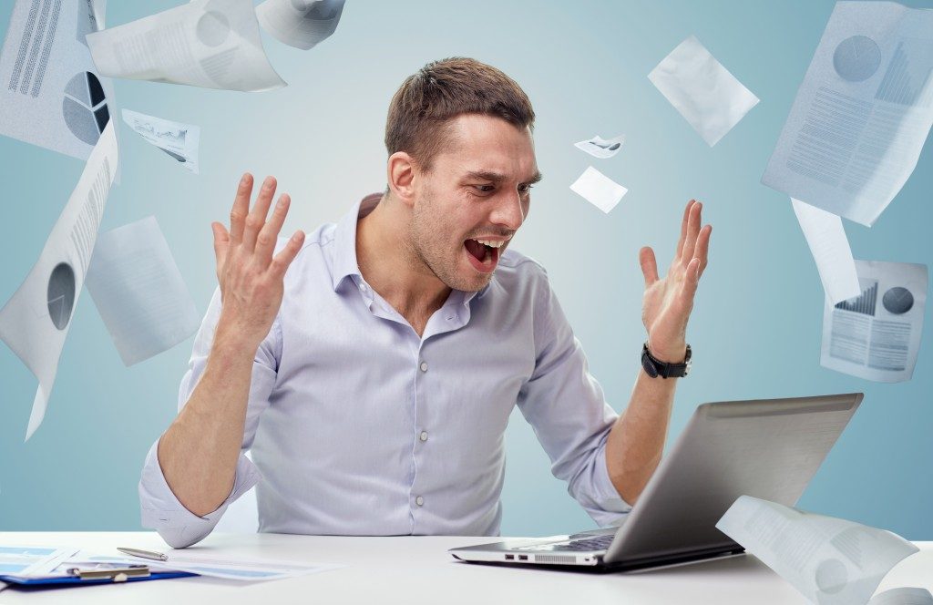 Angry businessman with laptop computer shouting over blue background and falling papers