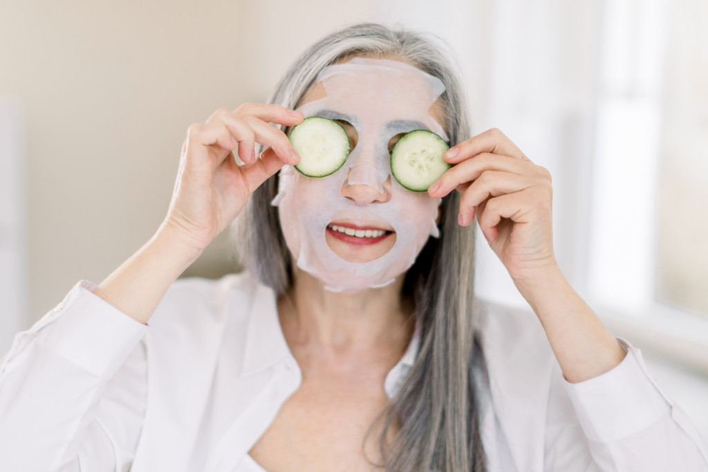 Smiling gray-haired senior woman at home, with facial sheet anti-aging mask on face skin, holding fresh cucumber slices, covering her eyes